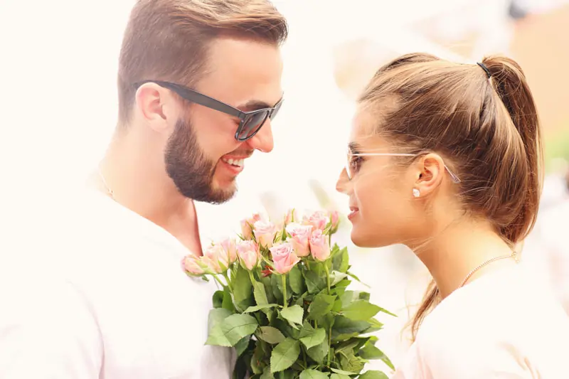 Ein Mann hält einen Strauß Rosen und lächelt seine Freundin an. Beide tragen Sonnenbrillen.