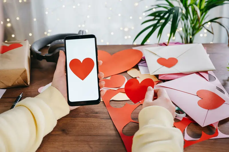 Ein Smartphone mit einem Herz-Emoji auf dem Bildschirm, gehalten von einer Person, umgeben von handgemachten Herzdekorationen und Briefen, ideal für WhatsApp Ich liebe dich Sprüche und Botschaften für deinen Schatz.