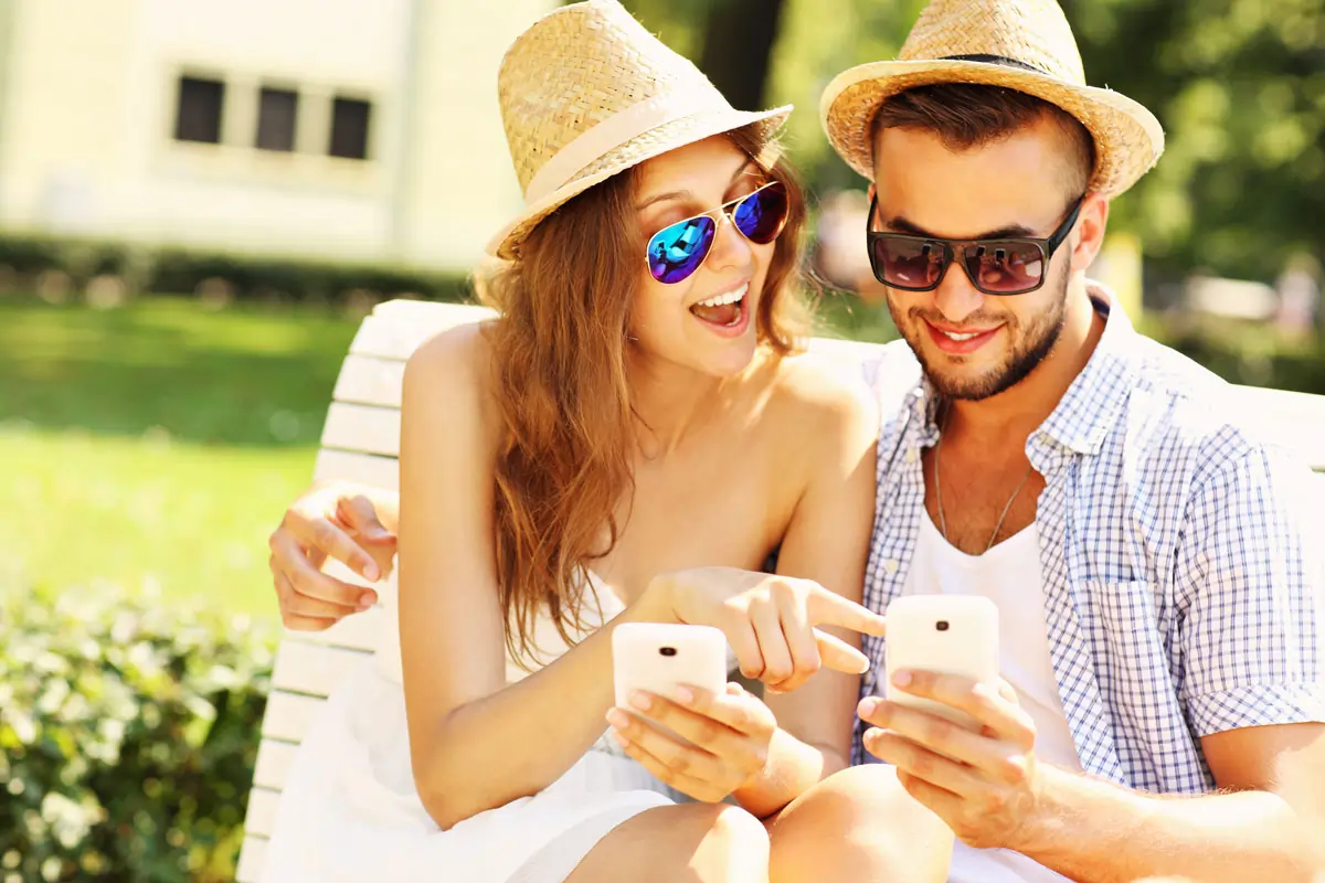 Ein glückliches Paar sitzt auf einer Bank im Park und schaut lächelnd auf ihre Smartphones, während sie WhatsApp Ich liebe dich Sprüche austauschen.