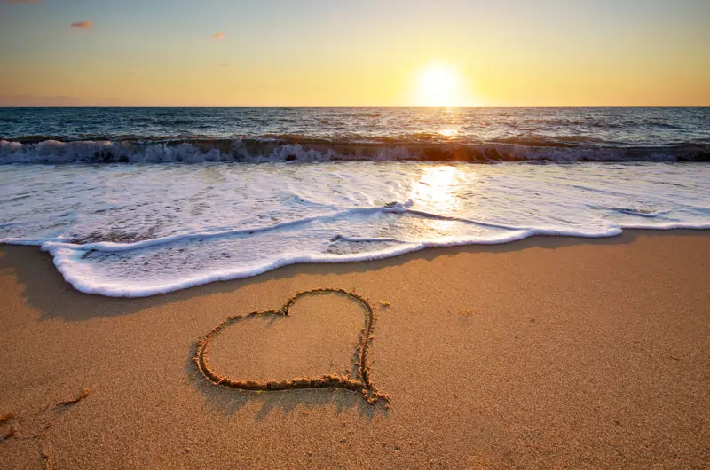 Ein Herz, das in den Sand am Strand gezeichnet wurde, wird langsam von den Wellen weggespült, was die Vergänglichkeit von Liebe zeigt.
