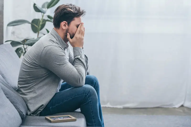 Mann sitzt verzweifelt auf dem Sofa mit den Händen vor dem Gesicht und einem Foto, das Eifersucht und emotionale Ausbrüche nach einer Trennung darstellt.