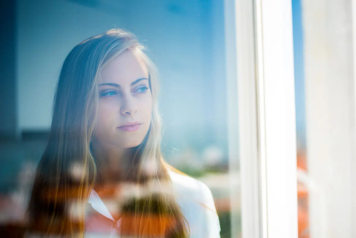 Nachdenkliche junge Frau schaut durch ein Fenster und reflektiert über ihre vergangene Beziehung.