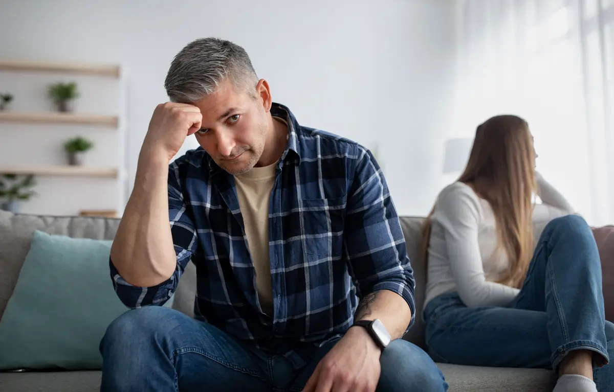 Ein nachdenklicher Mann sitzt bedrückt auf dem Sofa, während eine Frau im Hintergrund sitzt und sich abgewandt hat, was auf die emotionale Belastung nach einer Trennung oder dem Ende einer Affäre hinweist.